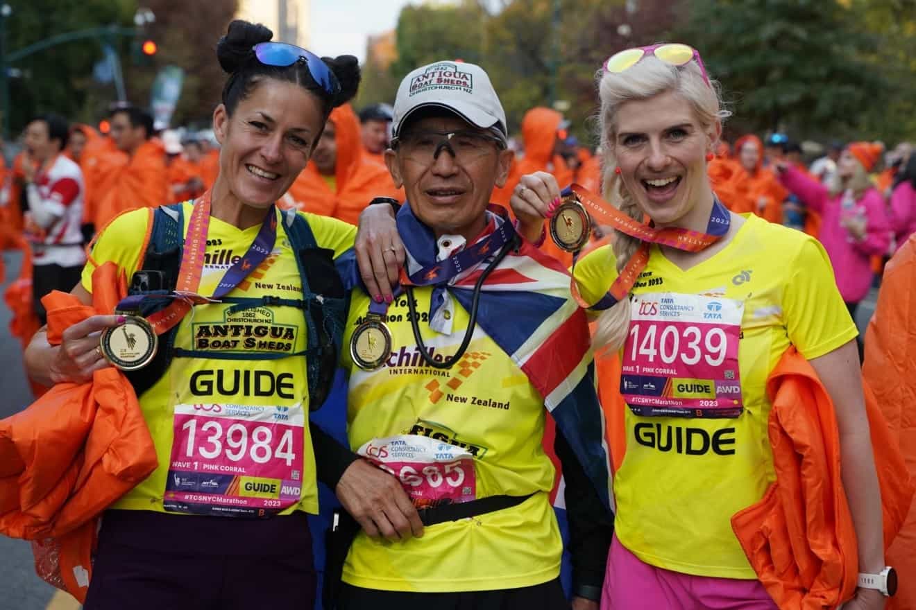 One Achilles athlete and two guides, one on either side, standing close to each other with the crowd in the background at the NYC Marathon