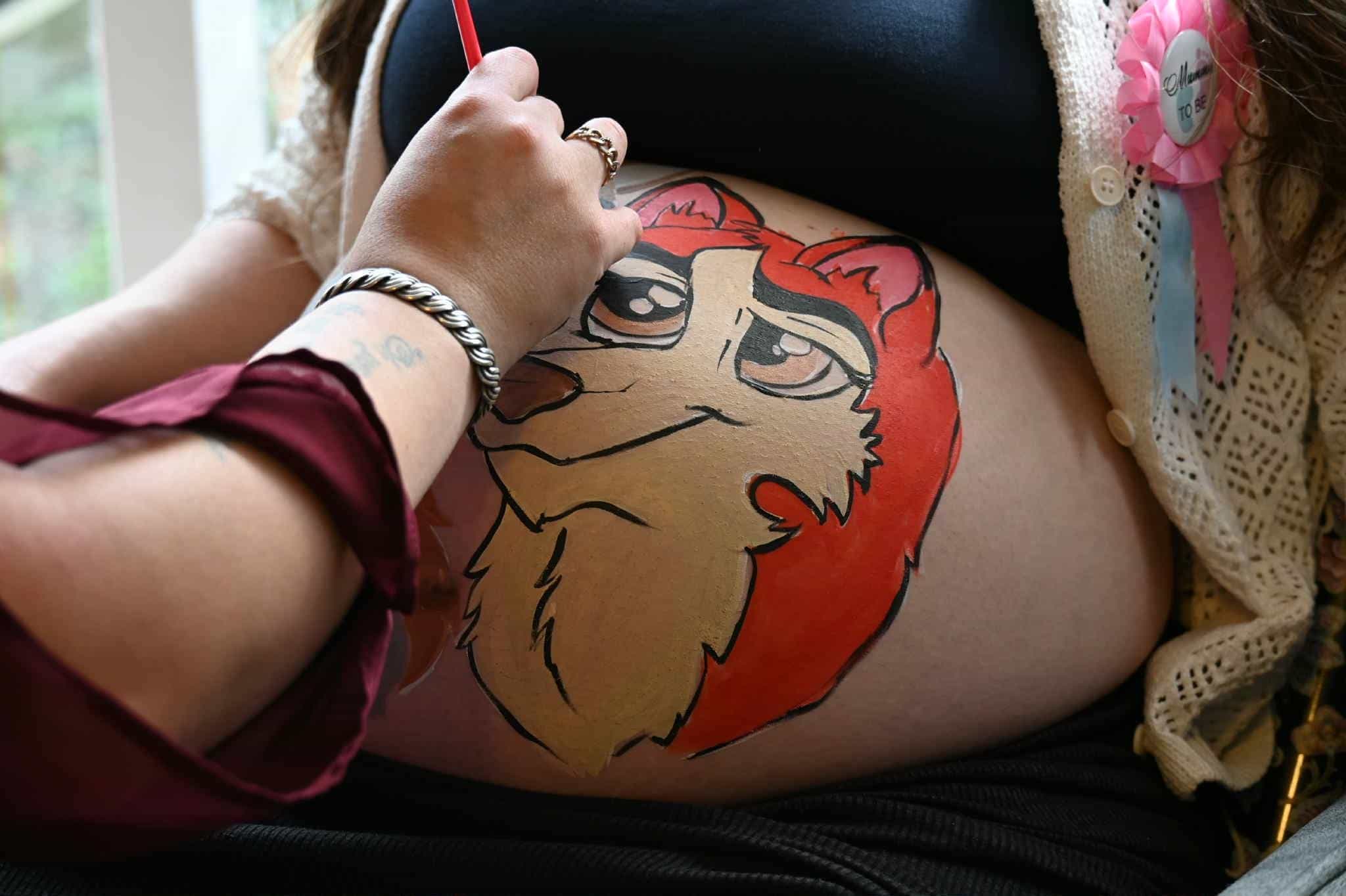 A close up of a baby bump in the process of having a smiling cartoon fox painted on it shades of orange and white. The artist's arms are just in view, wearing a crimson top and a pearl bracelet. 