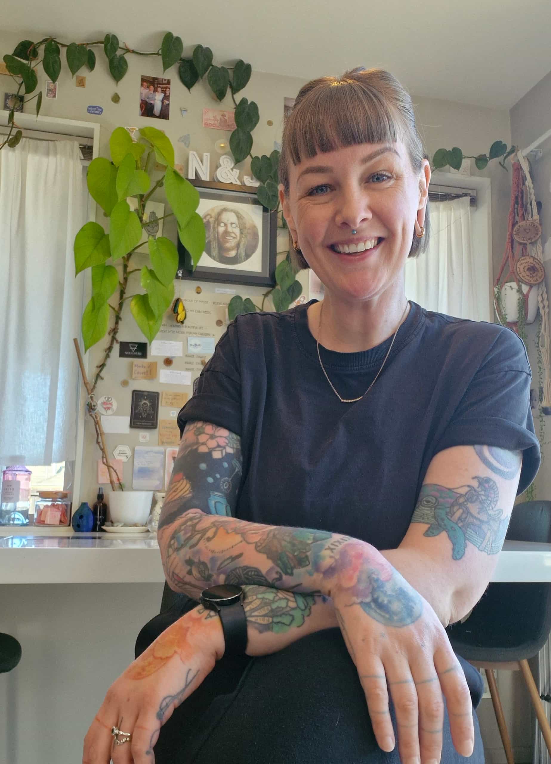A smiling woman with a bobbed hair cut, brightly coloured tattoos and a septum piercing wearing a black t shirt. There are houseplants and pictures in the background. 