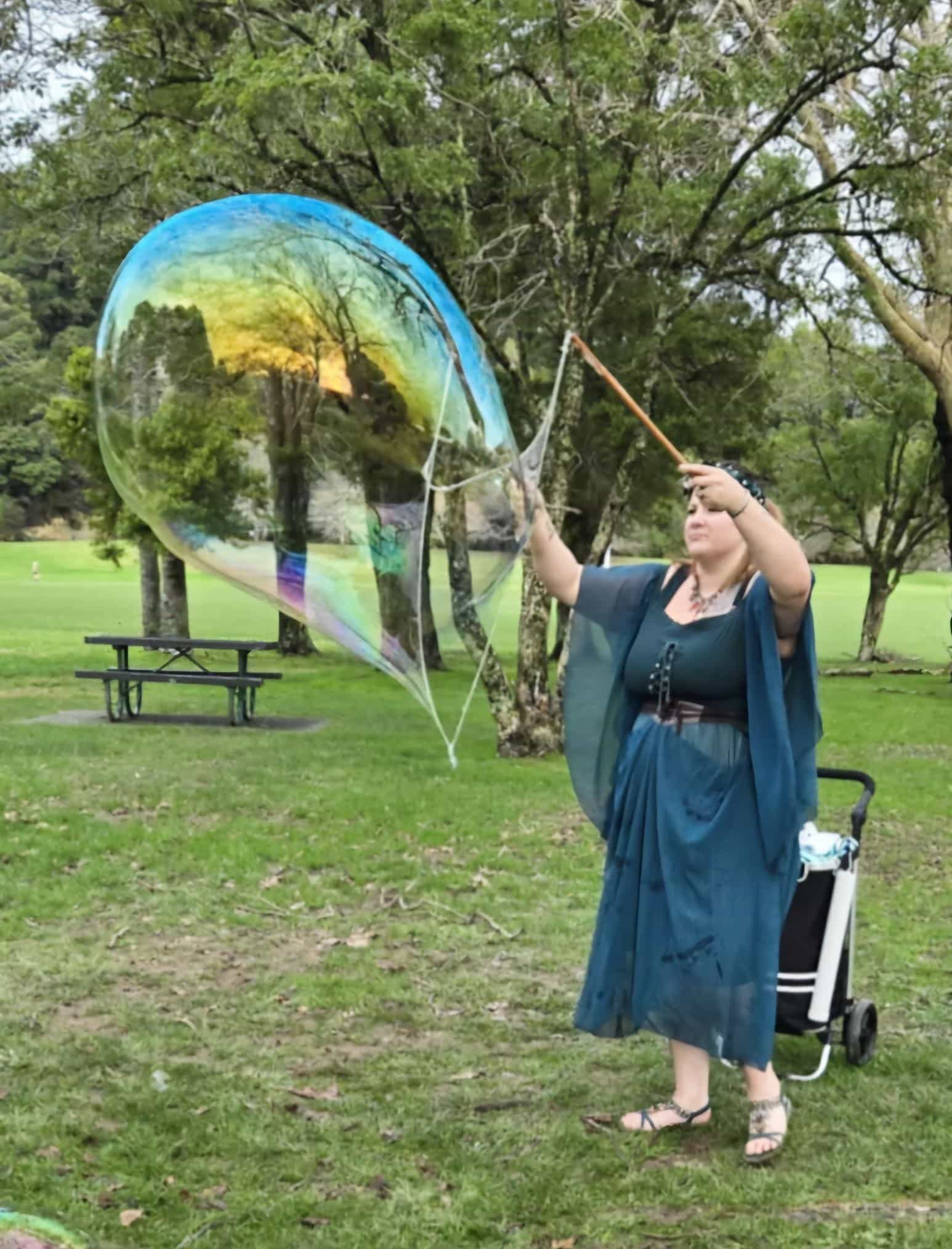 A woman wearing a blue dress holding a bubble wand with a bubble the same height as she is coming out of it. 