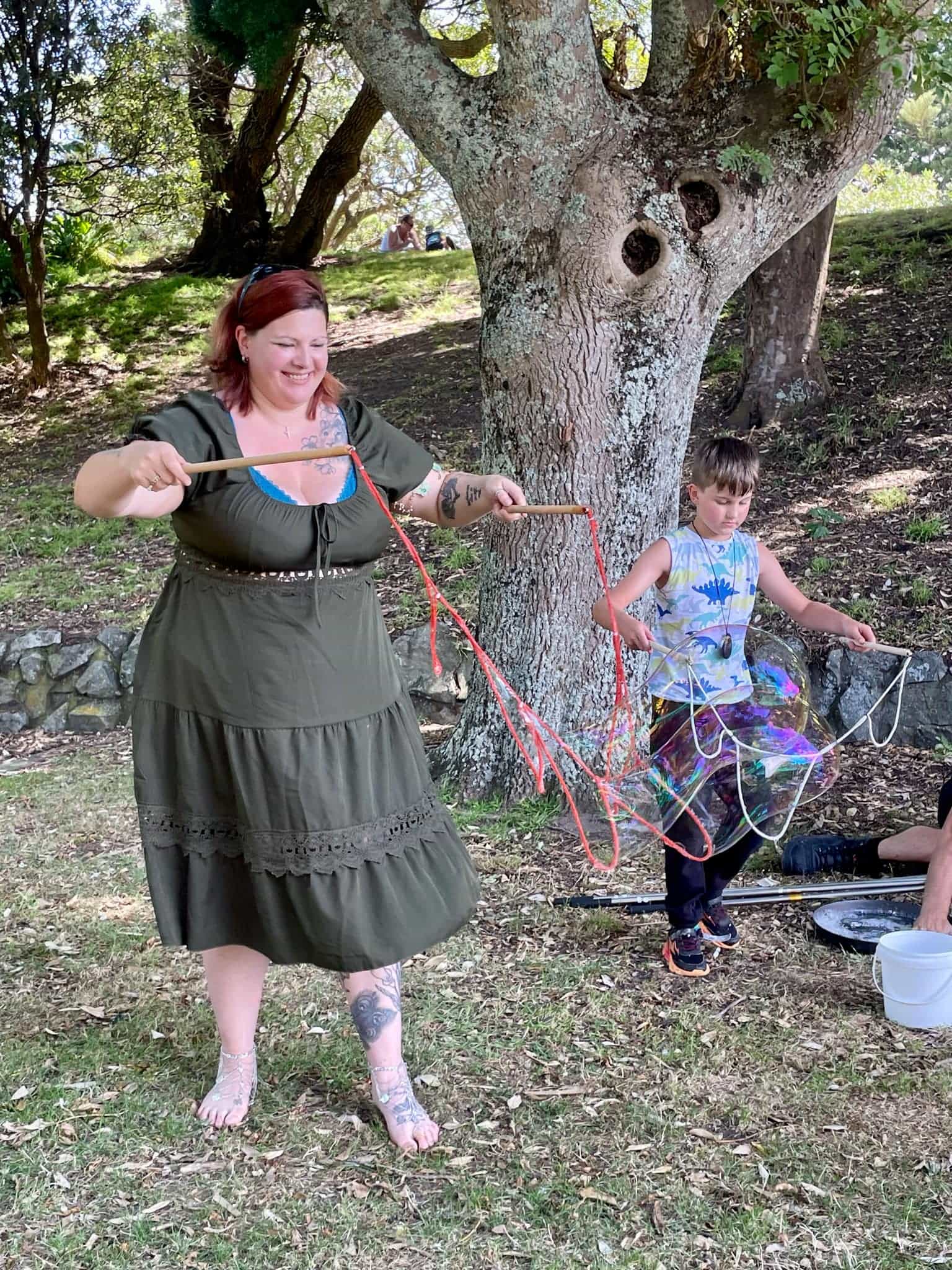 A red haired woman wearng a black sundress, smiling and standing barefoot on the grass. A boy is standing next to her wearing black trackpants and a blue tie-dyed singlet. Both are holding bubble nets. 