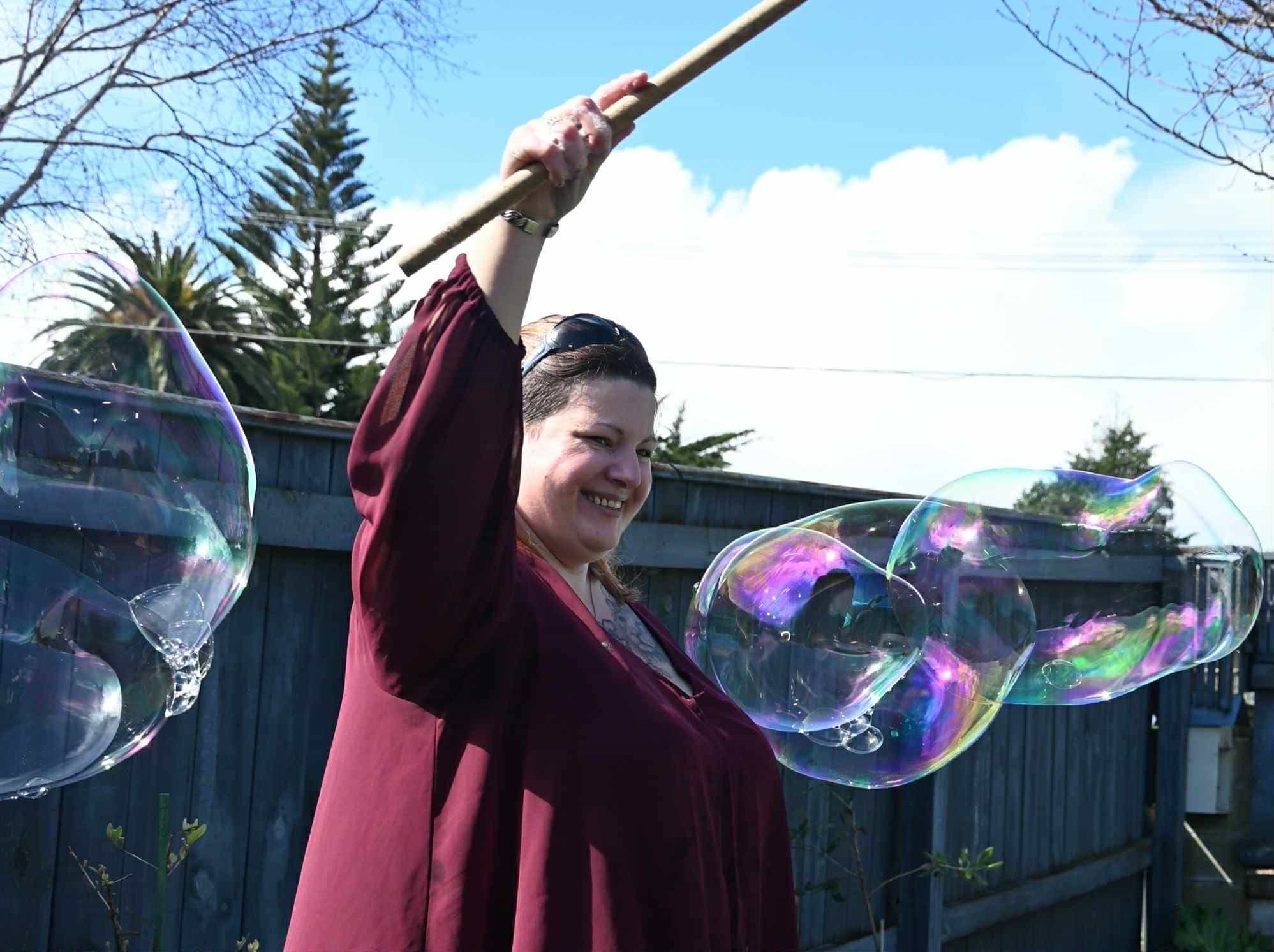 A woman wearing a crimson top surrounded by large bubbles. She is holding a bubble wand and smiling. 