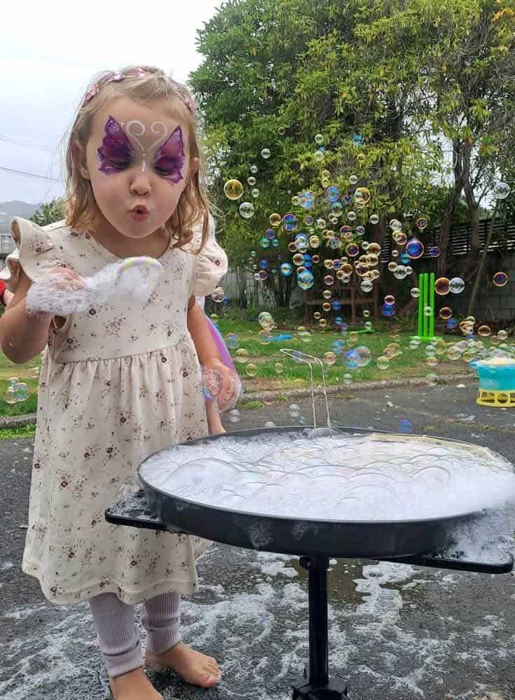 A toddler wearing a pale pink sundress, blowing bubbles while standing in front of a table of bubble mixture. She has a purple butterfly painted across her eyes.  