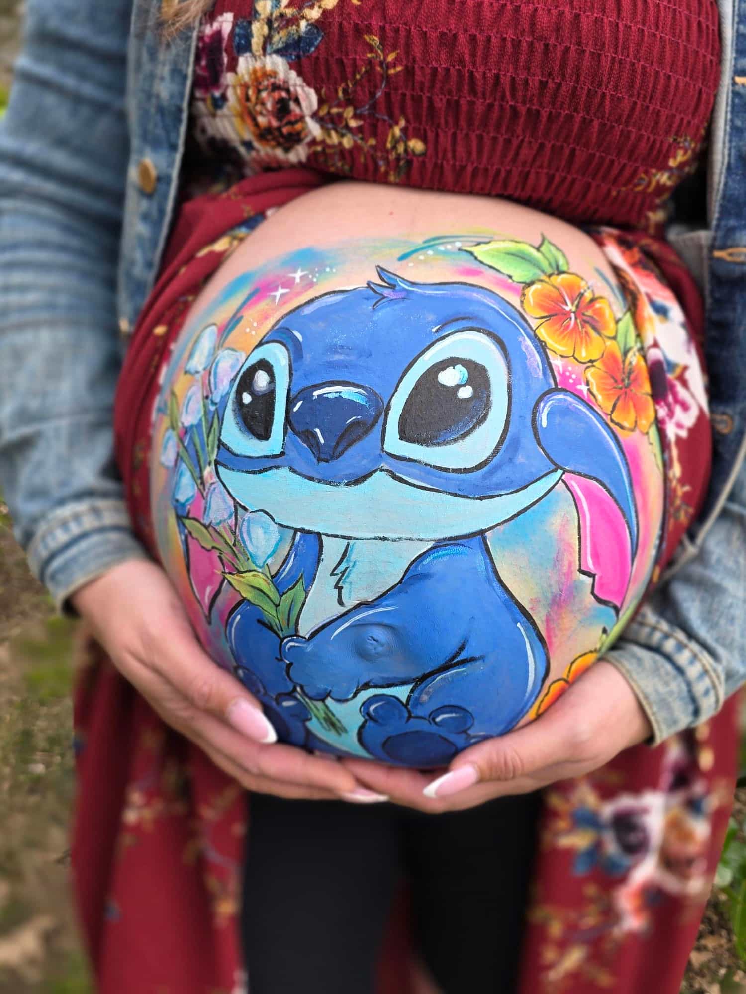 A close up of a woman wearing a crimson cotton dress, denim jacket and black leggings. The front of her dress is raised and she is cradling a baby bump with a blue cartoon creature (Lilo from Lilo and Stitch) painted on it, surrouned by tropical flowers. 