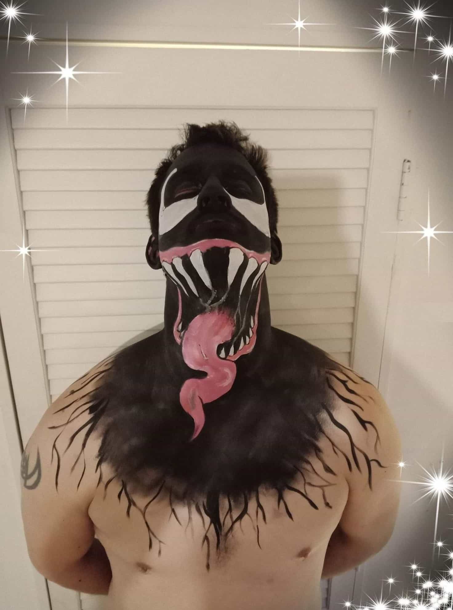 A shirtless man with the top half of his torso covered in black face paint, and his face painted black and white. He has his head tilted back to reveal sharp teeth and a long curly tongue painted on his neck, resembling a Mimic from Dungeons and Dragons. 