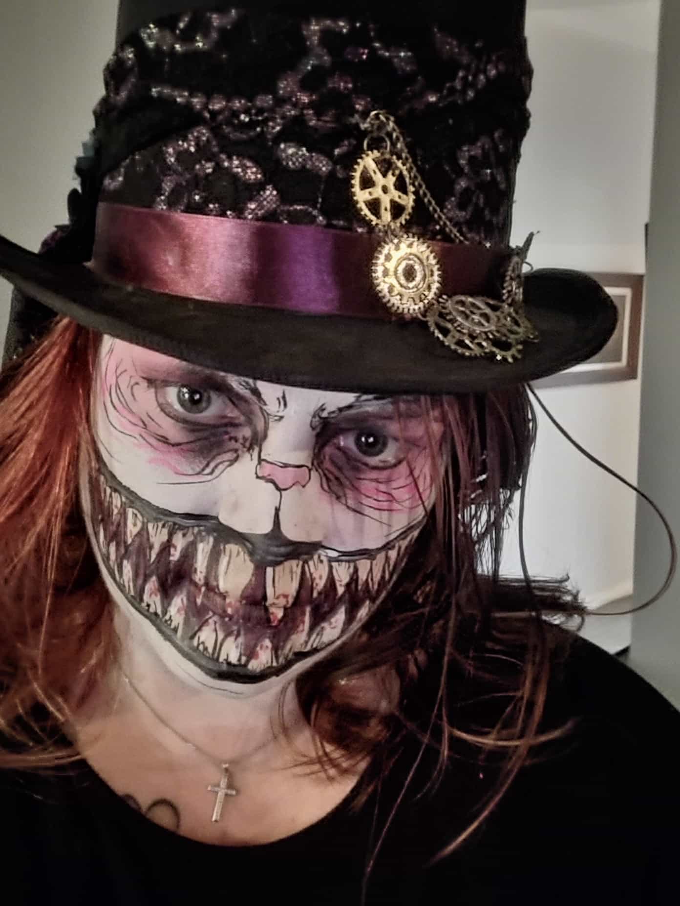 A woman (Christina) with her face painted to look like an evil Cheshire cat, with large distorted teeth right across her face. She is wearing a black top hat with a purple ribbon and clock fittings on it. 