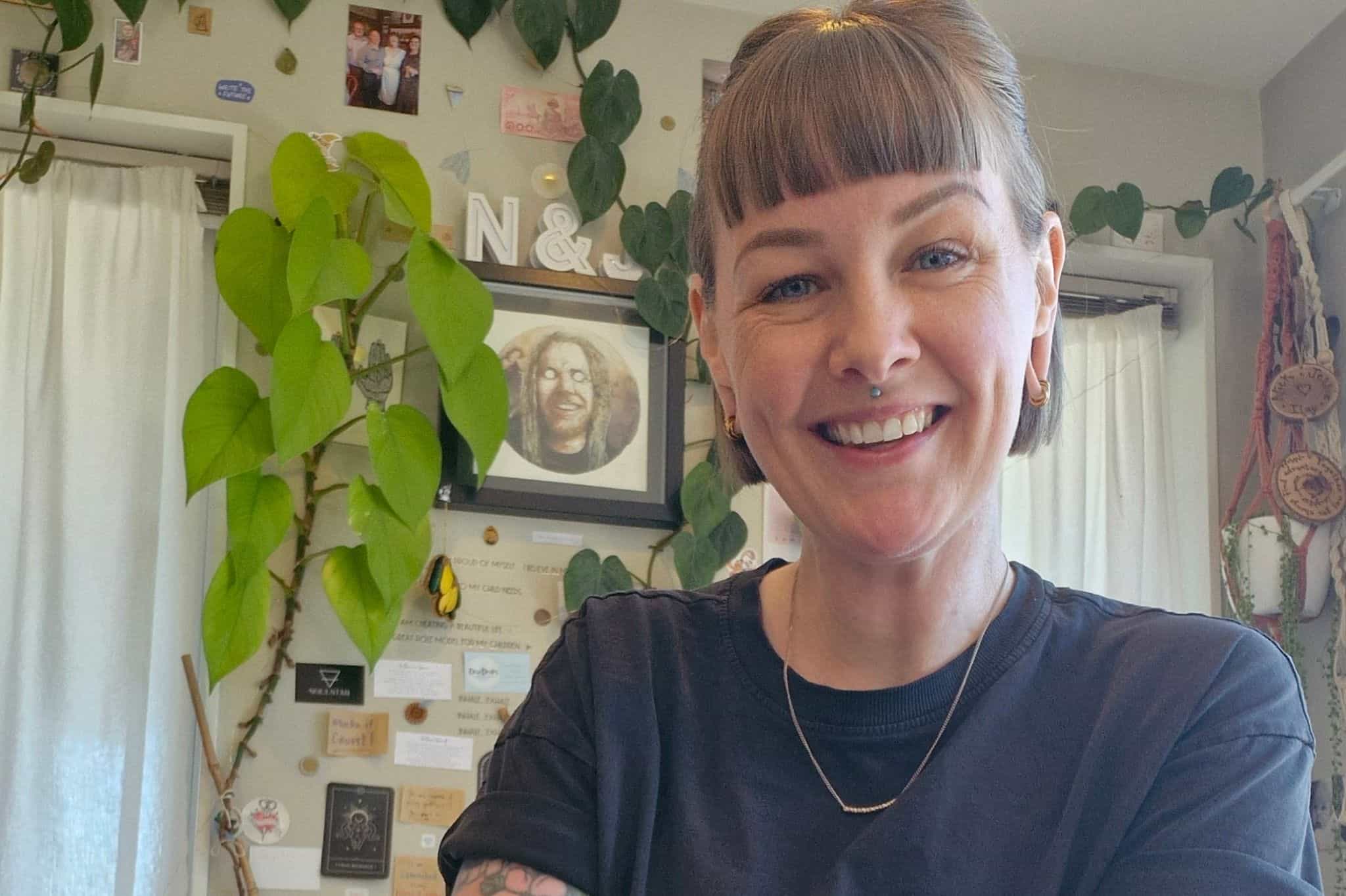 A smiling woman with a bobbed hair cut and a septum piercing wearing a black t shirt. There are houseplants and pictures in the background. 