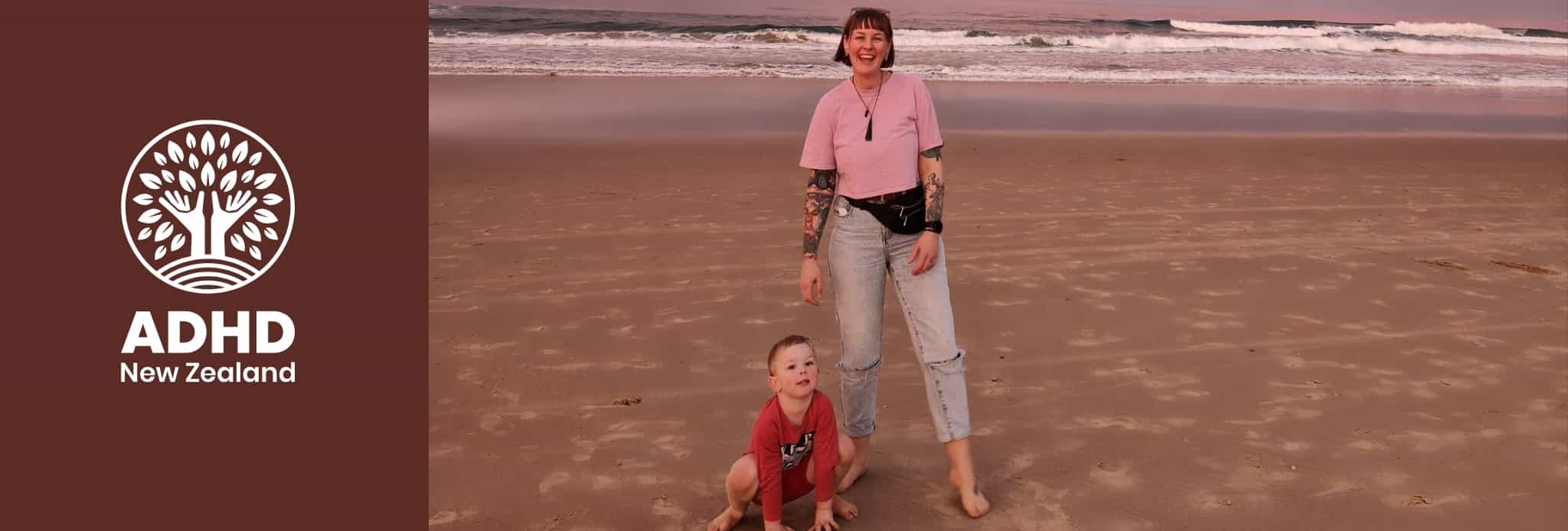 A smiling woman dressed in pink and a young boy wearing red on a beach. There are breaking waves and a pink and blue sunrise behind them. 