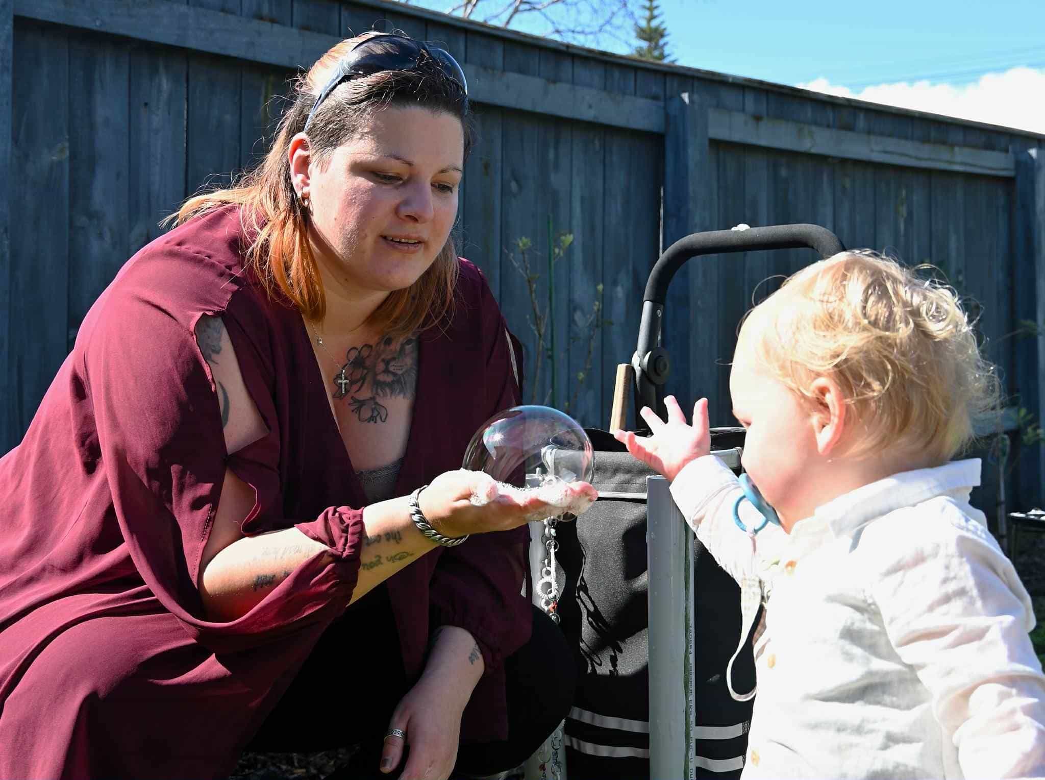 A woman wearing a crimson top, holding a bubble in her hand. She has crouched down to show it to a fascinated toddler. 
