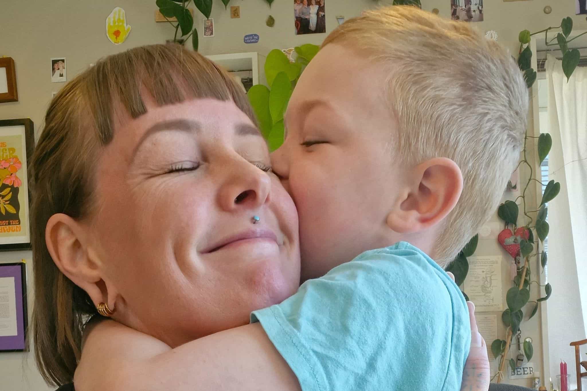 A woman with brown bobbed hair and a septum piercing hugging a small boy wearing a pale blue t shirt tightly. Both have their eyes scrunched closed and are smiling. 