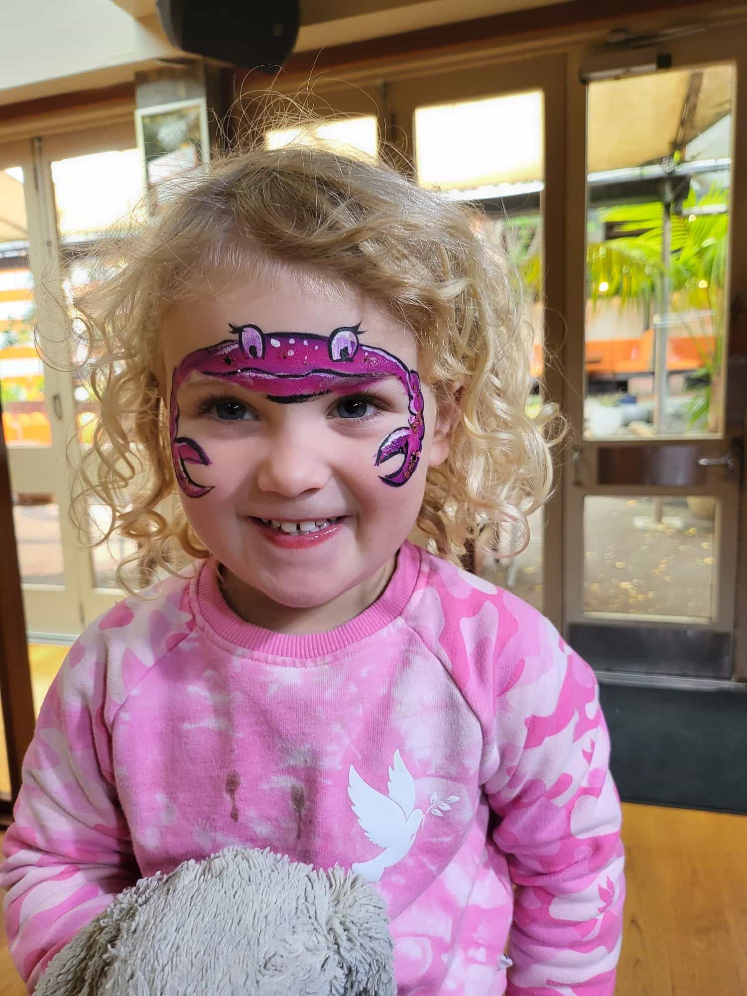 A young girl with a purple crab painted across her forehead. She is wearing a tie-dyed sweatshirt and showing two front teeth as she grins