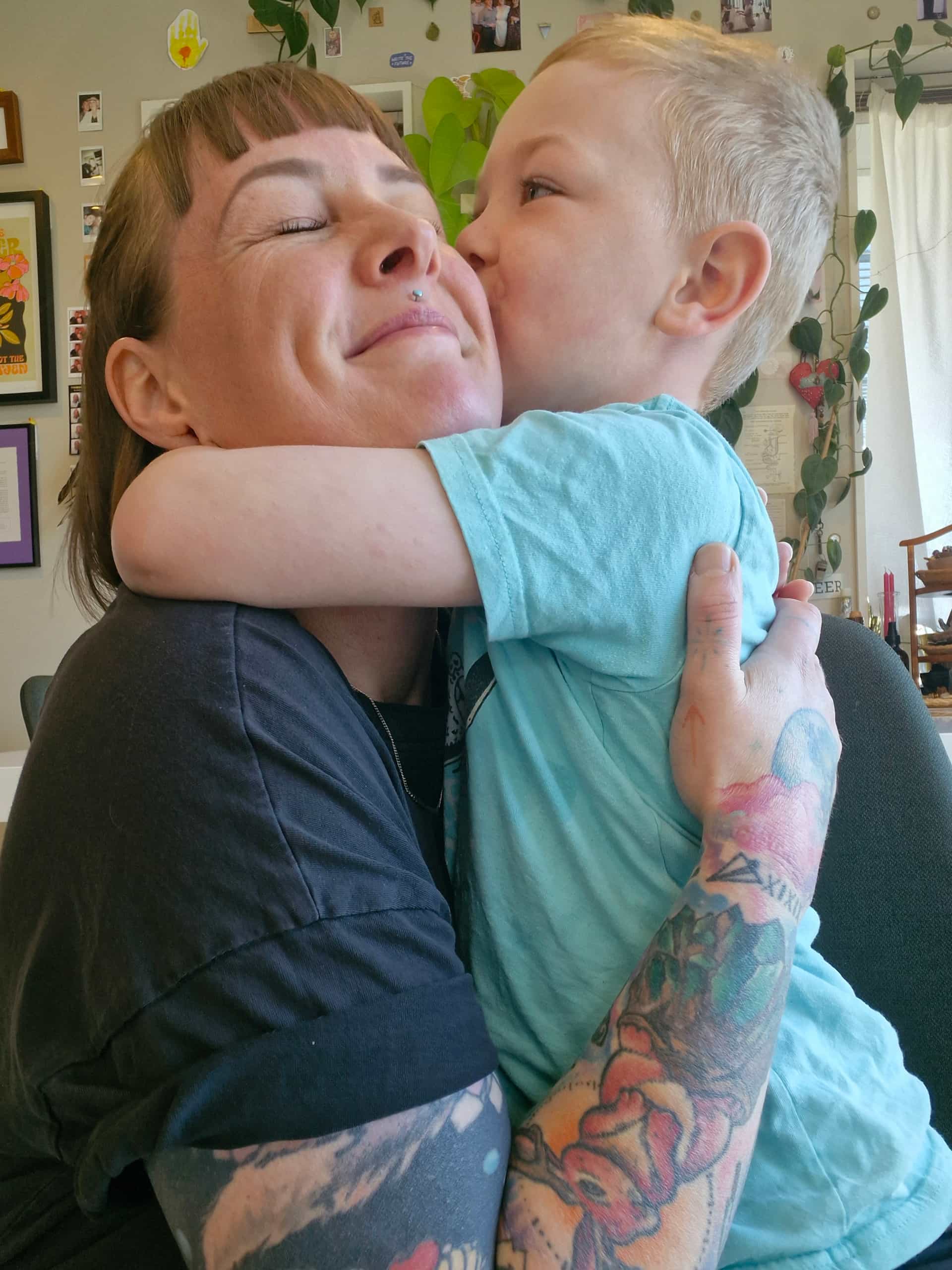 A woman with bobbed hair, bright coloured tattoos and a septum piercing hugging a small boy wearing a pale blue t shirt tightly. Both have their eyes scrunched closed and are smiling. 