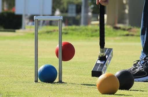 A grass level view of a jump shot in croquet, with the red ball attempting a jump over the blue ball that is lodged in the jaws of the hoop.